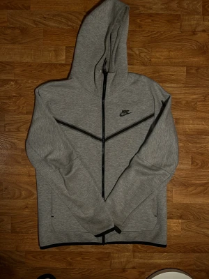 Grå Nike zip hoodie med huva M - Säljer en grå zip hoodie från Nike i storlek M. Jackan har hel dragkedja, huva och svarta detaljer som logga och dragkedja på ärmen. Tillverkad i mjuk bomullsblandning, perfekt för chill dagar eller när du vill ha en sportig look.