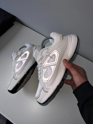 Dior B30 vita sneakers - Säljer ett par Dior B30 sneakers i vitt med grå och svarta detaljer. Skorna har en chunky sula, reflekterande paneler och stor Dior-logga på sidan. Tillverkade i mesh och skinn med snörning framtill. Riktigt snygga och trendiga sneakers för dig som vill sticka ut.