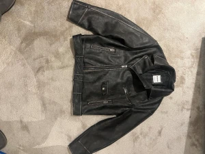 Svart skinnjacka från Bershka - Säljer en svart skinnjacka från Bershka med klassisk bikerstil. Jackan har flera dragkedjor, bred krage med tryckknappar och bälte i midjan. Snyggt detaljerad med synliga sömmar och en cool, lite matt finish. Perfekt för dig som vill ha en edgy look.