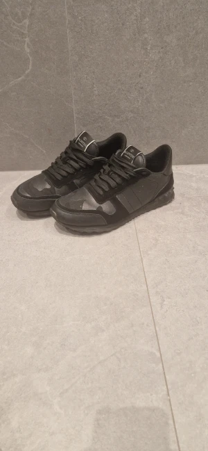 Valentino Garavani svarta sneakers - Svarta sneakers från Valentino Garavani med coolt kamouflagemönster och platt sula. Skorna har snörning, rund tå och detaljer i både mocka och skinn. Märkeslogga på plösen och insidan. Snygga nitar på hälen ger extra edge.