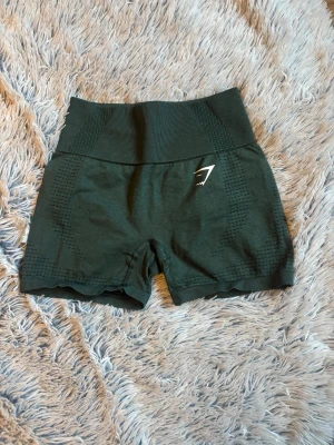 Mörkgröna Gymshark träningsshorts - Säljer ett par mörkgröna träningsshorts från Gymshark med hög midja och ventilerande mesh-detaljer längs sidorna. Stretchigt material som sitter tight och bekvämt, perfekt för gymmet eller löpning. Diskret Gymshark-logga framtill.