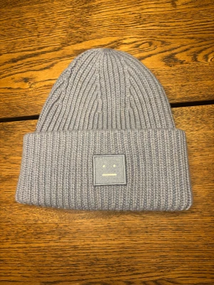 Ljusgrå Acne Studios mössa - Snygg ljusgrå ribbstickad mössa från Acne Studios med det klassiska Face-logotypen framtill. Mössan är i mjuk ull och har bred uppvikt kant för extra stilpoäng. Perfekt för kalla dagar och enkel att matcha med din jacka. Den är fläckig på baksidan där av pris