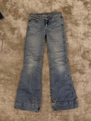Bootcut jeans från Wrangler, ljusblå - Bootcut jeans från Wrangler i ljusblå tvätt med klassiska fem fickor och bruna detaljer på bakfickan. Jeansen har breda benslut och snygga slitningar för en avslappnad vibe. Perfekta för dig som gillar retrostil och vill sticka ut.
