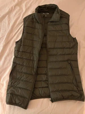 Mörkgrön dunväst från Ströva XS - Snygg mörkgrön dunväst från Ströva i storlek XS. Västen har quiltad design med horisontella sömmar, hög krage och dragkedja framtill. Två praktiska fickor med dragkedja på sidorna. Perfekt att slänga över en hoodie för extra värme.
