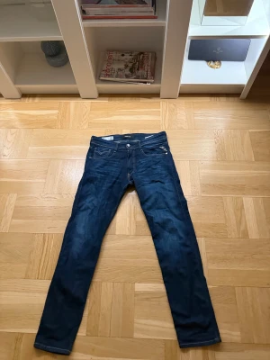 Replay Anbass mörkblå jeans - Snygga mörkblå Replay Anbass jeans med klassisk femficksdesign och smal passform. Jeansen har en stilren look med diskreta slitningar och detaljer som dragkedja och knappstängning. Perfekta för dig som gillar en modern och clean stil.strlek 31-32 som ny användas 2 gånger 