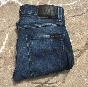 Mörkblå jeans från Nudie Jeans Co - Snygga mörkblå jeans från Nudie Jeans Co med klassisk femficksdesign och subtila slitningar. Jeansen har en rak passform och är tillverkade i slitstarkt denim med detaljerade sömmar och logotyp på bakfickan. Perfekt för dig som gillar stilrena och tidlösa jeans.
