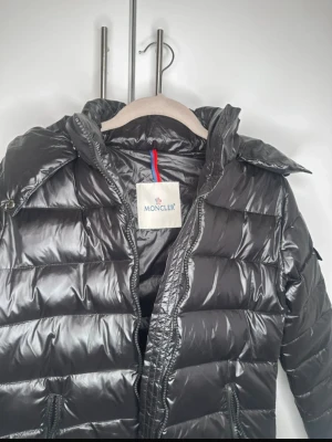 Svart Moncler pufferjacka med huva - Säljer en svart pufferjacka från Moncler med glansig finish och huva. Jackan har dragkedja framtill, två sidofickor med dragkedja och Moncler-logga på ärmen. Den är vadderad och har en modern, clean look som passar perfekt när det är kallt ute. Pris kan diskuteras vid snabb affär