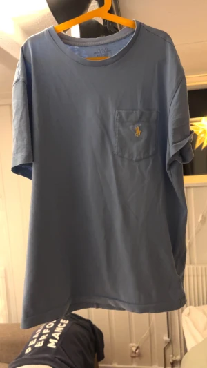 Blå t-shirt från Polo Ralph Lauren - Snygg blå t-shirt från Polo Ralph Lauren med klassisk gul broderad logga på bröstfickan. T-shirten har rund hals och korta ärmar. Perfekt för dig som gillar stilrena plagg.