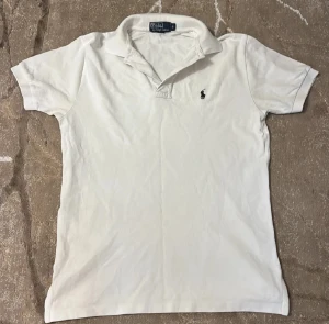 Vit pikétröja från Ralph Lauren - Klassisk vit pikétröja från Ralph Lauren i storlek S. Tröjan har korta ärmar, krage och knäppning vid halsen. Den är tillverkad i bomull och har den ikoniska broderade polospelaren på bröstet. Perfekt för en clean och stilren look.