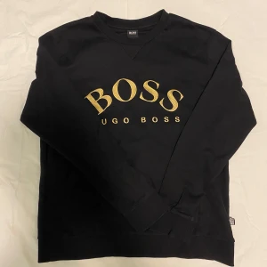 Svart tröja från Hugo Boss - Snygg svart tröja från Hugo Boss med guldfärgad logga på bröstet. Tröjan har en klassisk rund halsringning . Perfekt för en stilren och avslappnad look. Sitter som stor S eller M