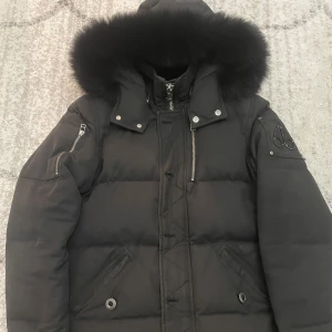 Svart dunjacka Moose Knuckles päls - Säljer en svart dunjacka från Moose Knuckles med fet pälskantad huva och snygga detaljer och har flera dragkedjor.Färgen har gått bort lite på olika delar av jackan men annars är den väldigt fin och bra.Perfekt för kalla vinterdagar.