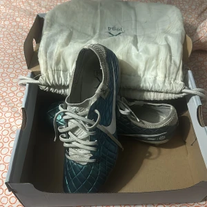 Nike X 1994 blå fotbollsskor - Säljer ett par Nike X 1994 fotbollsskor i metallicblått med vita detaljer och snörning. Skorna har ett rutmönstrat mönster på ovandelen och kommer med en vit tygpåse med dragkedja och tryck. Perfekta för dig som vill sticka ut på planen.