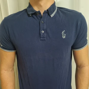 Marinblå pikétröja från Ralph Lauren - Snygg marinblå pikétröja från Ralph Lauren med vit broderad logga på bröstet och texten POLO/67 på ärmen. Tröjan har klassisk krage, knappar framtill och vita ränder på krage och ärmslut. 