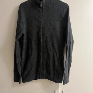 Mörkgrå kofta från Jack & Jones - Säljer en mörkgrå stickad kofta från Jack & Jones i storlek L. Koftan har hel dragkedja framtill, ribbade muddar och hög krage. Materialet är mjukt och passar perfekt till kyligare dagar. Enkel och stilren design som funkar till det mesta.