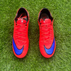 Röda Nike fotbollsskor med blå swoosh - Säljer ett par riktigt snygga röda Nike fotbollsskor med blå swoosh-detaljer. Skorna har snörning och är designade för att ge bra grepp på planen. Perfekta för dig som vill sticka ut på matchen och satsa på både stil och funktion.