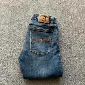 Blå jeans från Nudie Jeans Co, 28/32 - Snygga blå jeans från Nudie Jeans Co i klassisk tvätt med orange sömmar och ikonisk bakficka. Modellen har en smal passform och raka ben. Perfekta för dig som gillar stilrena och tidlösa jeans. Materialet är mjukt och slitstarkt denim.