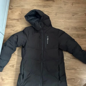 sail racing jacka - Svart dunjacka från sail racing  med huva och dragkedja. Jackan har en clean look, bröstficka med dragkedja och är vadderad med 500 FP dun från Allied Feather + Down. Perfekt för kalla dagar och har en stilren vibe. Varan är redo att hämtas eller fraktas. Köpt från Boozt för 3000 föra vintersäsongen, därför säljer jag jackan billigt.