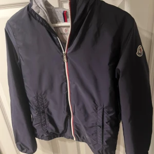 Mörkblå vindjacka från Moncler - Snygg mörkblå vindjacka från Moncler med grå huva och vit dragkedja med röd och blå detalj. Jackan har två fickor med knappar och klassisk Moncler-logga på ärmen. Perfekt för vår och höst, tillverkad i polyester och har en sportig vibe. Den är köpt från monclears hemsida och är knappt använd.😁 