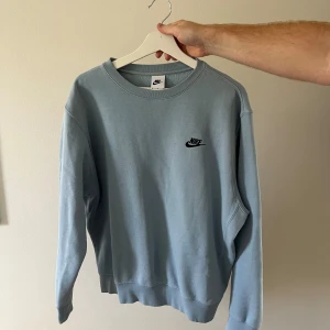 Ljusblå Nike sweatshirt i bomull M - Nike sweatshirt i ljusblå färg med svart broderad logga på bröstet. Tröjan har rund hals, långa ärmar och ribbade muddar vid ärmslut och nederkant. Materialet är 80% bomull och 20% polyester, vilket gör den mjuk och skön att bära.