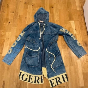 Blå parkajacka Tommy Hilfiger  - Unik blå parkajacka i jeansmaterial med huva, stora fickor och vita snören i midjan. Ärmarna har guldfärgade tryck och nederkanten har en bred beige kant med svart text. Perfekt statement-plagg för dig som vill sticka ut.