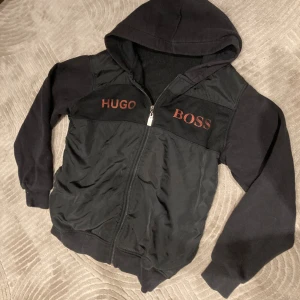 Svart hoodie från Hugo Boss med dragkedja - Svart hoodie från Hugo Boss med orange logga broderad på bröstet. Tröjan har huva, långa ärmar och hel dragkedja framtill. Materialmix med mjuka ärmar och vadderad framsida ger en sportig och stilren look,den passar dig om du är 160-165cm, Perfekt för dig som gillar streetwear.