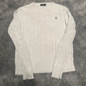 Vit kabelstickad tröja Polo Ralph Lauren - Vit kabelstickad tröja från Polo Ralph Lauren med klassisk rund halsringning och diskret broderad logga på bröstet. Tröjan är i bomull och har långa ärmar samt ribbade muddar för en snygg passform.