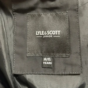 Svart pufferjacka Lyle & Scott Junior - Svart pufferjacka från Lyle & Scott Junior med huva och gul logotyp på ärmen. Jackan har quiltad design, dragkedja framtill och långa ärmar. Perfekt för kalla dagar och har en clean, stilren look som funkar till streetwear.