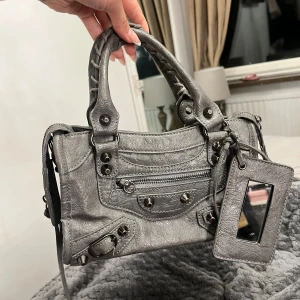  grå handväska i skinn - Snygg grå handväska i skinn med coola nitar, dragkedja framtill och ett matchande hängande spegel-fodral. Väskan har två flätade handtag och detaljerade metalldelar som ger en edgy vibe. Perfekt storlek för det viktigaste och riktigt trendig look.