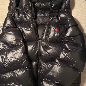 Svart pufferjacka från Polo Ralph Lauren - Snygg svart pufferjacka från Polo Ralph Lauren med glansig finish och röd broderad logga på bröstet. Igensydda hål på ryggen (se bild 5) absolut inget man märker eller ser. Pris kan diskuteras, alla frågor besvaras. GARANTERAR ÄKTHET, BEVIS FINNS