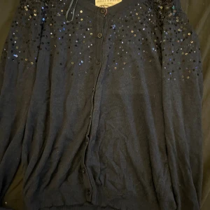 Mörkblå kofta med paljetter Holly & White - Snygg mörkblå kofta från Holly & White med paljettdetaljer över axlar och bröst. Koftan har knappar framtill och är långärmad, perfekt för att lyfta en enkel outfit. Mjuk och skön känsla.