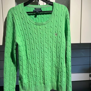 Grön kabelstickad tröja från GANT - Fräsch grön kabelstickad tröja från GANT i stretchig bomull. Tröjan har rund hals, ribbade muddar och ett litet broderat GANT-emblem på bröstet. Perfekt för dig som gillar klassisk stil med en färgklick.