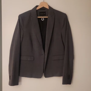 Grå kavaj från STOCKH LM, storlek 42 - Snygg grå kavaj från STOCKH LM i klassisk modell med två fickor fram och slät finish. Tillverkad i 95% ull och 5% elastan med fodrad insida i polyester och viskos. Perfekt för en stilren look och passar till många olika outfits.