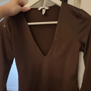 långärmad v-ringad body - Säljer en brun långärmad topp från H&M i storlek S. Toppen har en djup v-ringning och är figurnära med en clean, enkel design. Materialet är mjukt och stretchigt, perfekt för att styla med jeans eller kjol. Snygg och tidlös färg som funkar året runt. Perfekt till fest 