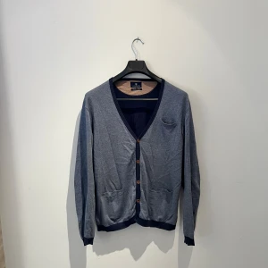  Gråblå Carshmere kofta från Hansen & Jacob - Kofta i cashmereblandning från Hansen & Jacob. Gråblå med mörkblå detaljer, v-ringad och knäppning med bruna knappar. Två fickor framtill och snygga armbågslappar i mörkblått på ärmarna. Perfekt för dig som gillar stilrena och mjuka plagg. Hör av er vid frågor eller fler bilder