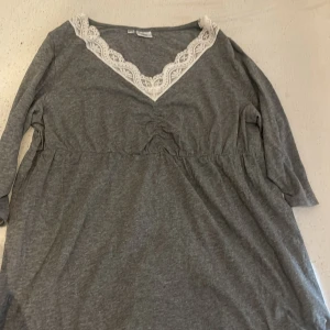 Grå klänning med spets från bpc - Supersöt grå klänning från bpc med vit spets runt v-ringningen. Klänningen har trekvartsärm och är gjord i mjuk bomull. Den har en avslappnad passform och snygga rynkdetaljer under bysten. Perfekt för dig som gillar enkel stil 