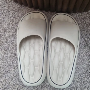 Vita chunky sandaler med platt sula - Säljer ett par vita chunky sandaler med bred rem över foten och platt sula. Sulan har ett mönster som påminner om bikake-struktur och en tunn svart rand längs kanten. Perfekta för sommardagar och chill hemma.