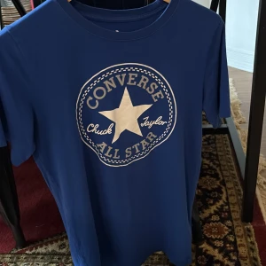 Blå Converse All Star t-shirt L - Snygg blå t-shirt från Converse med det klassiska All Star-trycket i vitt framtill. Rund halsringning och korta ärmar. Perfekt för dig som gillar streetstyle och vill ha en ikonisk look. Storlek L i barnstorlek men passar xs-s