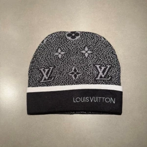 Svart LV mössa - Snygg svart och grå mössa från Louis Vuitton med klassiskt LV-mönster och vit kant. Mössan är stickad och har tydlig Louis Vuitton-logga både fram och bak. Perfekt för dig som vill ha en lyxig och stilren accessoar i vinter.