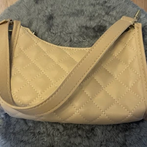 Beige quiltad axelväska  - Snygg beige axelväska med quiltat rutmönster. Väskan har en enkel, stilren design och stängs med dragkedja. Perfekt storlek för det viktigaste och passar till många olika outfits.