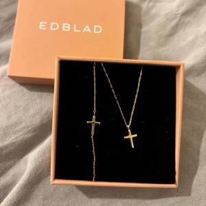 Guldigt korshalsband & armband från Edblad - Snyggt halsband & armband från Edblad med tunn kedja i guldton och ett stilrent kors som hänge. Perfekt för dig som gillar minimalistiska smycken med en personlig touch. Levereras i original ask. Nypris för halsbandet: 299 Nypris för armbandet 249