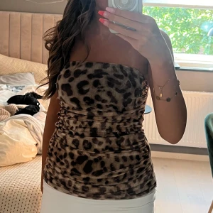 Leopardmönstrad bandeau tubtopp - Säljer en snygg bandeau tubtopp med leopardmönster i bruna och svarta toner. Toppen är åtsittande och har en lätt rynkad effekt längs sidorna. Perfekt för dig som vill sticka ut med ett djurmönstrat plagg. Materialet känns mjukt och stretchigt.