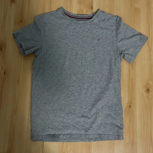 Grå t-shirt från Tommy Hilfiger - Snygg grå t-shirt från Tommy Hilfiger med klassisk logga broderad på bröstet. T-shirten har rund halsringning med en cool detalj i form av röd, vit och marinblå rand på insidan av kragen. Tillverkad i mjuk bomull och har en normal passform.