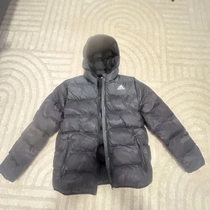 Svart pufferjacka från Adidas - Svart pufferjacka från Adidas med huva och dragkedja framtill. Jackan har två sidofickor med dragkedja och diskret Adidas-logga på bröstet. Perfekt för kalla dagar med sin vadderade design och sportiga look. 