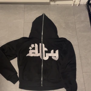 Svart hoodie Divide The Youth - Svart hoodie med hel dragkedja och stor huva från Divide The Youth. Framsidan har ett stort tryck i vitt med stjärndetaljer och baksidan har texten 'DIVIDE THE YOUTH' i vitt. Mjuk och skön, perfekt för streetwear-stil. Knappt använd.