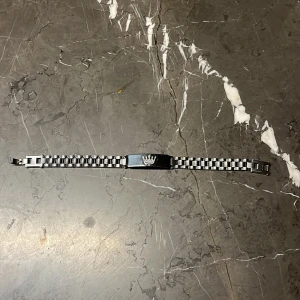 Silverfärgat armband med krona - Snyggt armband i silverfärgad metall med rutigt mönster och en platta med en graverad krona i mitten. Länken har en klassisk design som passar till många stilar och är lätt att justera med spännet.