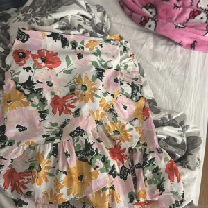 Blommig volangkjol från ONLY - Superfin blommig kjol från ONLY med volangkant och knyt i midjan. Kjolen har färger i rosa, gul, röd, grön och svart på vit botten. Perfekt för dig som gillar färg och mönster! Modellen är kort och har en flowy passform.