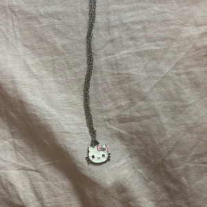 Hello Kitty halsband med kedja - Sött halsband med en silverfärgad kedja och en Hello Kitty-berlock. Berlocken är vit med svarta detaljer och har en rosa rosett på huvudet. Perfekt för dig som gillar kawaii och vill ha något gulligt och personligt.