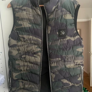 Camouflage dunväst från lagency  - Snygg dunväst i camouflage från Lagency med quiltad design och dragkedja framtill. Västen har två fickor med dragkedja och en patch med logga på bröstet. Perfekt för lager-på-lager och streetstyle.