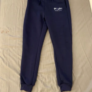 Trapstar - Snygga mörkblå mjukisbyxor från Trapstar med tryck på vänster lår och texten 'IT'S A SECRET'. Byxorna har snörning i midjan och ribbade muddar vid bensluten. Perfekta för chill eller streetwear-stil.