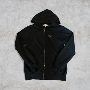Svart hoodie Comme des Garçons Play S - Svart hoodie från Comme des Garçons Play i storlek S. Tröjan har dragkedja, huva och fickor framtill. Ikoniskt hjärta med ögon broderat på bröstet och stort tryck på ryggen. Tillverkad i mjuk bomull, perfekt för en chill och stilren look.